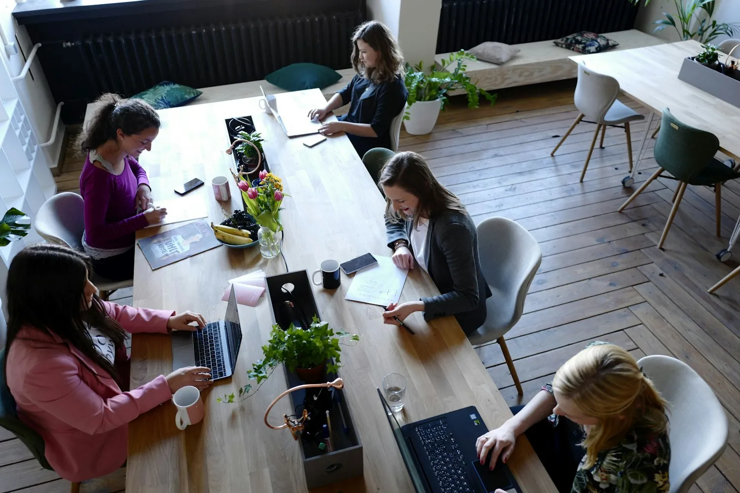 Plusieurs professionnels installés autour d'une grande table en bois dans un espace de travail partagé, ordinateurs portables ouverts